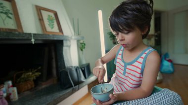 Evde oturan küçük bir çocuk, küçük bir kaseden kaşıkla yemek yiyor. Çocuk yemek yerken kameraya bakıyor, rahat bir ev ortamıyla çevrili.