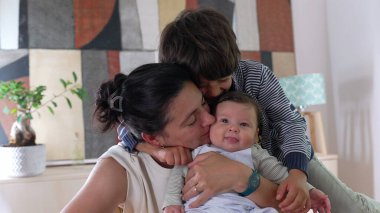 Anne ve büyük kardeş, bebeği kucaklıyor iç mekanda iç açıcı bir aile bağları var. Anne-çocuk ve kardeş evde sevgi ve şefkatle kaynaşıyor.