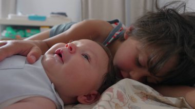 Büyük çocuk bir yatağın üzerinde bebeğin yanında yatıyor, bebeğe şefkatle bakarken bebeğin kafasına hafifçe dokunuyor. Kardeşler arasındaki kapalı bağ anı