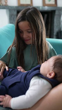 Kanepede oturan genç bir kız kucağındaki bebeğe sevgi dolu bir şekilde bakıyor, bebeğin elini nazikçe tutuyor. Evde şefkatli bir kardeş anı, sıcaklığı, aile bağını ve sevgiyi yakalıyor.