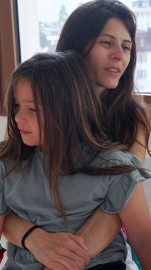 Anne pencerenin kenarında otururken küçük kızını sevgiyle kucaklıyor. Her ikisi de sakin ve sevgi dolu bir anı paylaşıyor, bir yuva ortamında besleyici bağı ve sıcaklığı sergiliyor.