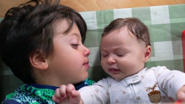 Küçük çocuk, bebek kardeşinin yanında uzanıyor. Bebek elinde oyuncak tutarken dikkatle bakıyor. Bağlanma anını gösteriyor. Kardeşler bir paspasın üzerinde uzanıyorlar.