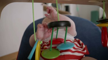 Bebek bir fedaide uzanıyor, küçük ellerle renkli asılı oyuncaklara uzanıyor, dokuları keşfediyor, oyuncu duyusal gelişim gösteriyor, odak ve merak gösteriyor.