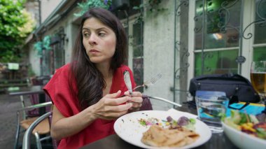 Dışarıda yemek yiyen bir kadın tabağından çatal dolusu salata çıkarmaya odaklanmış. O, bir restoranın açık verandasında otururken yemeğin tadını çıkarırken, meşgul görünüyor.