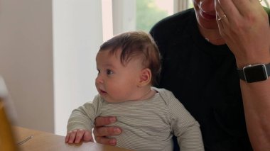 Bebek yemek masasında annesinin kucağında oturuyor, kameranın dışında bir şeye tuhaf bir şekilde bakıyor. Annenin eli nazikçe bebeği destekliyor.