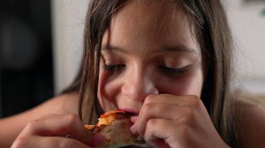 Bir parça pizza kabuğunu ısıran genç bir kız, iki eliyle tutuyor, zevk anında gözlerini kapatıyor, bir masada oturuyor, yüz ifadesine odaklanıyor.