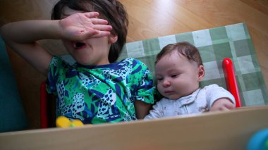 Genç bir çocuk yere uzanıyor, burnunu eliyle siliyor, burnu akan bir burundan dolayı, sakin görünen bir bebeğin yanında, sessiz oyun zamanı boyunca bir kardeşlik anı.