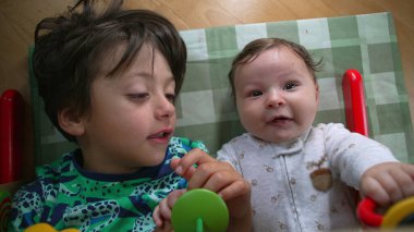 Genç bir çocuk ve bebek yerde yatıyor, birbirlerine bakıyorlar, sevinçle gülümseyen bebek, neşeli ve neşeli bir kardeşlik bağı kurma anını vurguluyorlar.