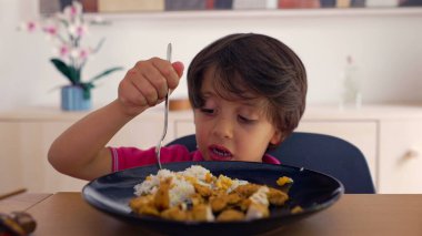 Genç çocuk çatalla yemek yemeye, sakin bir evde yemek ve pilav yemeye, yemek saatinde konsantrasyon ve bağlılık göstermeye odaklanmış.