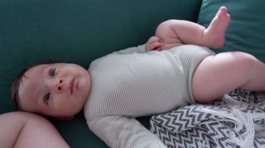 Bebek kanepede yatar, sakin ve meraklı bir ifadeyle yukarı bakar. Rahat bir ev ortamında bebeğin rahatlamış davranışlarını yansıtan huzurlu bir an.