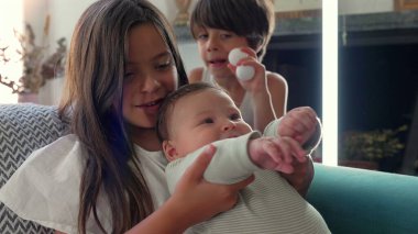 Kanepede otururken kucağında bebek olan genç bir kız ve arka planda izleyen bir oğlan. Rahat bir ev ortamında kardeşlik bağı ve aile birliği.