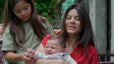 Annesi ikisini de kucağında tutarken, küçük kız kardeşini şefkatle teselli ediyor. Bebek üzgün görünüyor ve aile açık hava ortamında şefkatli bir anı paylaşıyor.