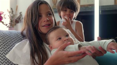 Kanepede otururken kucağında bebek olan genç bir kız ve arka planda izleyen bir oğlan. Rahat bir ev ortamında kardeşlik bağı ve aile birliği.