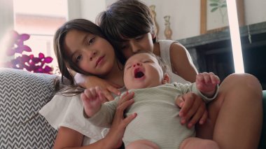 Kız bebeği kucaklarken bir oğlan yan taraftan izliyor, hepsi kanepede oturuyor. Sahne, huzurlu bir oturma odasında kardeş sevgisini ve aile bağını vurguluyor.