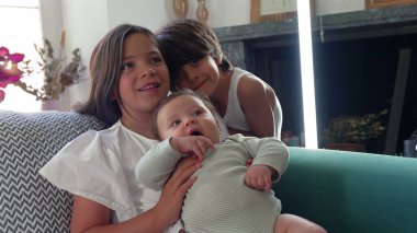 Bir oğlan arka plandan eğilip kanepede otururken bir bebeği kucağına alan genç bir kız. Sahne, kardeş sevgisini ve sıcak bir aile anını tasvir ediyor.