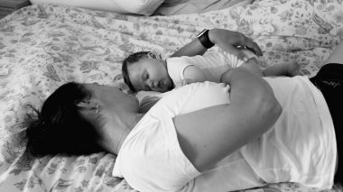 Anne, bebeği göğsünde huzur içinde uyurken yatakta dinleniyor. Sakin ve samimi bir aile bağını besliyor. Tek renkli, siyah ve beyaz bir evde.