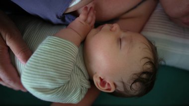 Bakıcının kollarında yatarken huzur içinde uyuyan bebek, bebeğin huzurlu yüz ifadesini ve kıvrık ellerini gösteren yakın çekim görüntüsüyle.