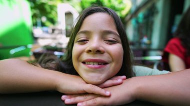 Çenesi ellerinin üzerinde dinlenmiş genç bir kız ışıl ışıl gülümsüyor. Neşeli, açık hava manzarası, mutluluk ve çocukluk masumiyeti.