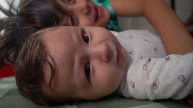 Bebeğin yerde yattığı yakın plan, gözleri sonuna kadar açık, arka planda kısmen görünen bir kardeş, bir bebek merakı anı ve erken kardeşlik etkileşimi