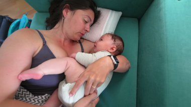 Anne kanepeye uzanıp bebeğini kucağında tutuyor, bebek merakla yukarı bakıyor, sıcak ve besleyici bir atmosfer yaratıyor.