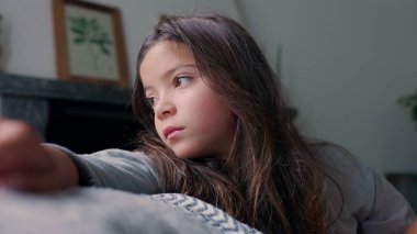 Kolunu dinlendirirken dikkatlice yan tarafa bakan genç kız. Sakin bir yansıma ve tefekkür anı, huzurlu bir ev ortamında çocukluk duygularının derinliğini yakalıyor.