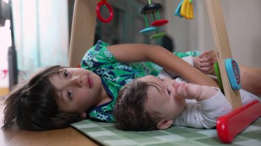 Genç bir çocuk oyuncak kemerinin altında bebekle şakalaşıyor, ikisi de yerde yatıyor, kardeş bağlantısı, neşe ve çocukluk zamanının ortak deneyimi