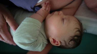 Bakıcının kollarında yatarken huzur içinde uyuyan bebek, bebeğin huzurlu yüz ifadesini ve kıvrık ellerini gösteren yakın çekim görüntüsüyle.