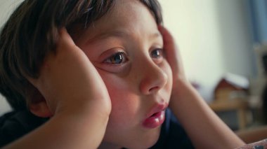 Genç çocuk ellerini yanaklarının üzerinde dinlendiriyor, odaklanmış ve özenli bir ifadeyle ekranı kapalı izliyor. Yakın çekim merakını ve konsantrasyonunu içeride yakalar.