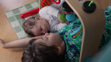 Küçük çocuk kardeşinin yanında yatıyor, oyuncakla şakalaşıyor. Bebek, büyük kardeşe bakıyor. Sıcak bir evde, ahşap bir spor salonunun altında birbirlerine bağlanırken.