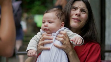 Anne tarafından tutulan bebek, açık bir kafede kucağında otururken merakla etrafına bakıyor. Bebek sakin ve memnun görünüyor, çevreyi geniş gözlerle keşfediyor. 