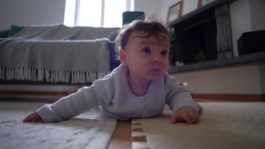 Bebek yerde yatıyor, meraklı bir ifadeyle bekliyor, ağırlığı kollarıyla tutuyor, emeklemenin ilk aşamaları, rahat bir yuva kurma, meraklı ve odaklanmış.