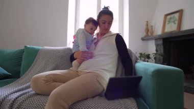 Anne kanepede bebeği tutuyor, ikisi de tablete bakıyor, birlikte ekran başında vakit geçiriyorlar, sıcak bir oturma odasında rahat bir aile zamanı geçiriyorlar.