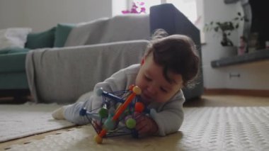 Bebek paspasın üzerinde yatıyor, iki eliyle renkli oyuncakları tutuyor ve güvenli bir ev ortamında erken gelişim sırasında şekilleri ve dokuları keşfediyor.