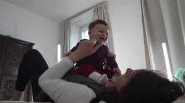 Anne bebekle yatakta uzanıyor, nazikçe oynuyor ve çocukla bağ kuruyor, erken duygusal gelişimi ve sıcak bir ev ortamında bağlantıyı besliyor.