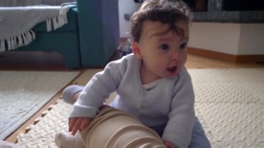 Bebek yerde oturup annesinin bacağını tutuyor, neşeli ve meraklı bir ifade, kapalı alanda aile bağları kurma anı, bebek keşfi çevresi, sakin bir evde neşeli etkileşim