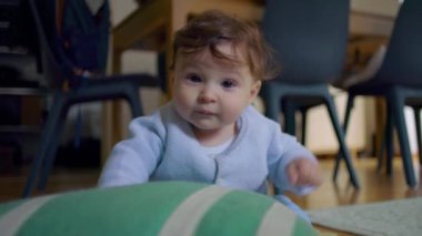 Bebek yerde oturuyor, tuhaf bir ifadeyle yukarı bakıyor, sıcak bir ev ortamında destek için ellerini kullanıyor, tamamen gözlem altında.