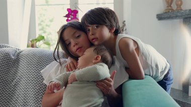 Bir oğlan kanepede otururken kucağında bebek tutan genç bir kız. Sahne, sıcak bir ev ortamında aile sıcaklığını vurgulayan hassas bir kardeş anı yakalıyor.