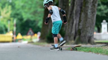 Küçük çocuk ağaçlarla kaplı bir patikada kaykay yaparken manevra yaparken kollarını denge için uzatmıştı. Kask ve koruyucu giysiler giyiyor. Odaklanma ve kararlılık gösteriyor.