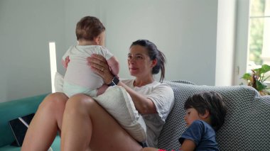 Anne kucağında bebeği tutuyor, büyük çocuk yanında dinlenirken onlara sevgiyle bakıyor, evdeki aile hayatının sıcaklığını ve yakınlığını gösteriyor.