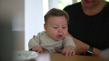 Bebek yemek masasında annesinin kucağında oturuyor, meraktan yana bakıyor. Annenin elleri nazik bir destek sağlıyor.