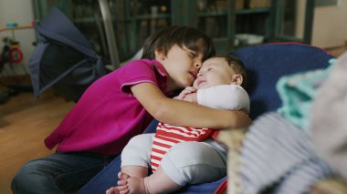 Büyük kardeş fedaide otururken bebeğe sarılıyor, sıcak bir ev ortamında sıcak ve şefkatli bir kucaklaşma sergiliyor, bebek merakla ve rahatlıkla kardeşine bakıyor.