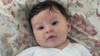 Çiçek desenli bir yatakta uzanmış bebek yukarı bakıyor, ağzı açık ve anlamlı gözleri var. Bebek çevresiyle meşgul görünüyor, sıcak ve doğal bir an yaratıyor.