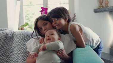 Bir oğlan kanepede otururken kucağında bebek tutan genç bir kız. Sahne, sıcak bir ev ortamında aile sıcaklığını vurgulayan hassas bir kardeş anı yakalıyor.