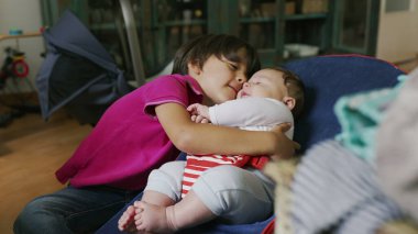 Büyük kardeş fedaide otururken bebeğe sarılıyor, sıcak bir ev ortamında sıcak ve şefkatli bir kucaklaşma sergiliyor, bebek merakla ve rahatlıkla kardeşine bakıyor.