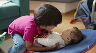 Küçük bir çocuk, bir fedainin içinde bebeğe yaslanıyor. İki kardeş sıcak bir ev ortamında bir anlık etkileşimi paylaşırken ilgi ve merak gösteriyor.