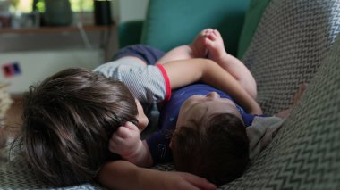 İki genç kardeş bir kanepede uzanıyor, birbirlerine sarılıyorlar, sıcak bir ev ortamında yakınlık ve yakınlık gösteriyorlar, doğal ışık odayı dolduruyor.