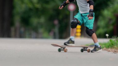 Genç bir kaykaycının yakın çekimi, bacaklarını ve koruyucu pedlerini gösteriyor. Kendini asfalt yolda ileri itmek için aktif olarak yerden kalkıyor.