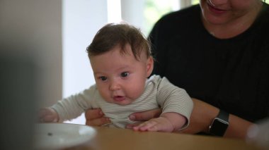 Bebek yemek masasında annesinin kucağında oturuyor, meraktan yana bakıyor. Annenin elleri nazik bir destek sağlıyor.