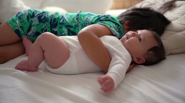 Büyük kardeş yatakta kucak kucağa yatıyor. Bebek gülümsüyor ve yukarı bakıyor. Kardeşi yanında yatarken sıcaklık ve sevgi gösteriyor.