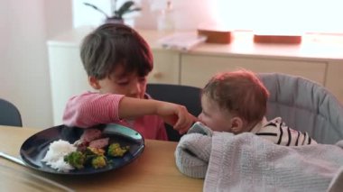 Küçük çocuk aile yemeği sırasında bebeğe uzanıyor, nazik bir jest, kardeş bağı, yemek masasında paylaşılan anlar, çocuklar arasındaki bağ ve bakım, sıcak atmosfer.